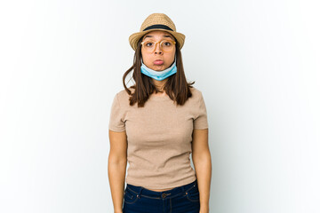 Young latin woman wearing hat and mask to protect from covid isolated on white background shrugs shoulders and open eyes confused.