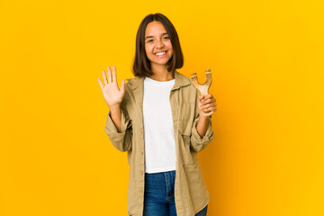 Young hispanic woman holding a slingshot smiling cheerful showing number five with fingers.