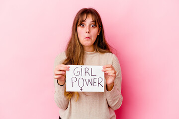 Young caucasian woman holding a girl power placard isolated on pink background shrugs shoulders and open eyes confused.