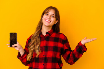 Young caucasian woman holding phone isolated on yellow background showing a copy space on a palm and holding another hand on waist.