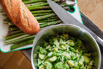 Asparagus on a plate with a knife, a baguette, and a bowl of  chopped brussel sprouts.
