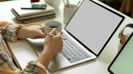 Fototapeta premium Side view of female hands holding coffee cup while working with laptop on office desk
