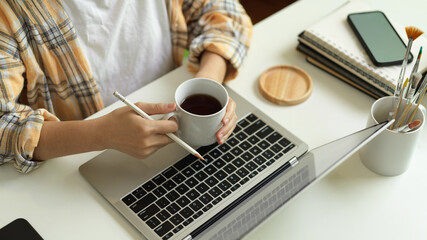 Fototapeta na wymiar Female freelancer hands holding coffee cup while working with laptop on home office desk