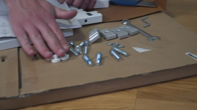 Young Caucasian man assembles white furniture from flat box using instructions. Male bought table online DIY during quarantine covid 19. Lockdown coronavirus ordering furniture home and self-assembly