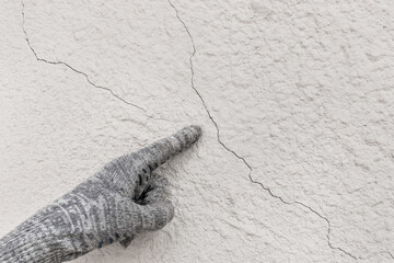Hand of industrial worker in construction glove points to cracks on white plaster wall texture background