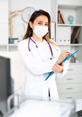 Young female therapist in a medical mask, working in a clinic, fills out a patient's treatment appointment sheet on a ..tablet in the office