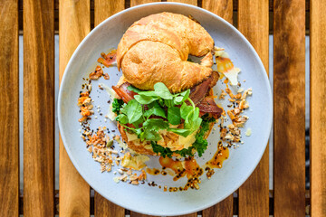 Scrambled Egg Croissant Burger with bacon, herbs and greens on a round plate with a  wooden table in the background.