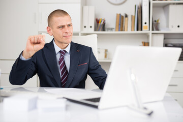 Positive european businessman having video call on laptop in modern office and holding fist