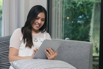 Pretty smiling young woman sitting on sofa and working on digital tablet.
