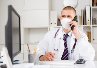 Male doctor in a protective mask, working in the clinic, consults a patient on the phone at a desk in the ..resident's office