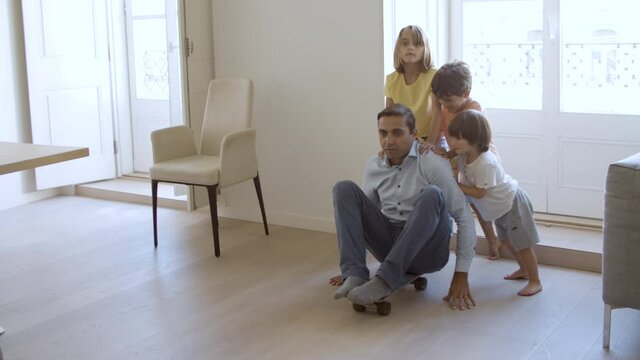 Kids and daddy having fun at home. Sibling children wheeling longboard with dad in living room, shouting, falling and laughing. Childhood or parenthood concept