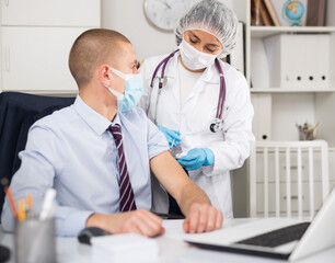 Doctor in face mask uses hypodermic needle and a syringe to put a shot of drug to patient in hospital office