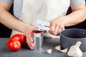 Obraz na płótnie Canvas A woman is carefully opening a can of tomato paste on a kitchen counter using a white plastic can opener. She is preparing a meal for which she uses both fresh and pureed preserved tomatoes