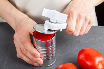 A woman is carefully opening a can of tomato paste on a kitchen counter using a white plastic can...