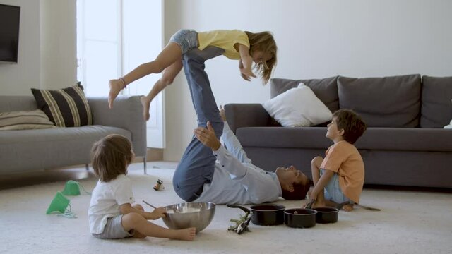 Father holding daughter on legs and lying on carpet. Happy Caucasian children playing in living room with dad. Two cute boys sitting on floor. Hand-held camera. Childhood and holiday concept
