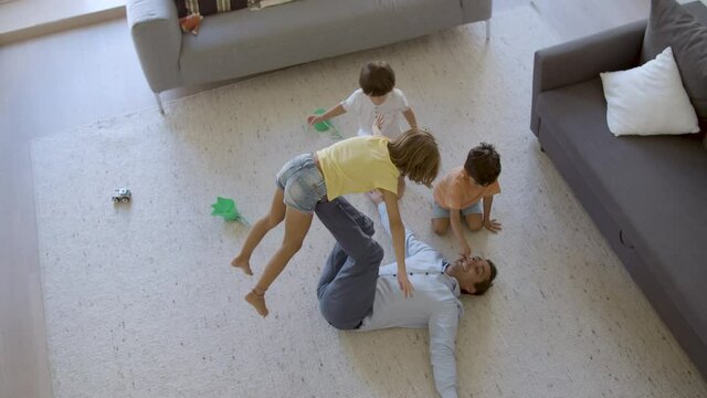 Smiling dad holding girl on legs and lying on carpet. Happy cute kids having fun in living room with father. Cute kids playing together. Static camera. Top view. Childhood and weekend concept