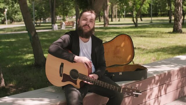 Tracking portrait shot of male street musician with beard sitting on long backless bench in park and playing song on guitar, then looking at camera