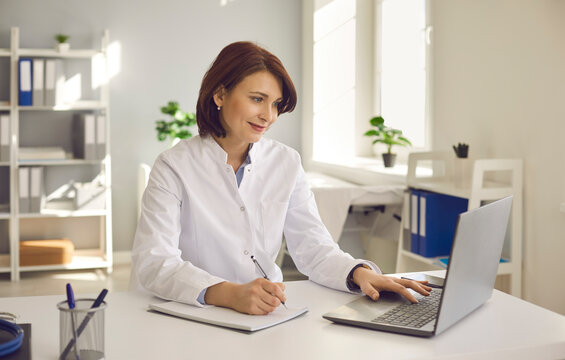 Female doctor writes notes while watching an online medical webinar or training seminar while sitting with a laptop in the workplace. Positive doctor does his best to provide quality medical care