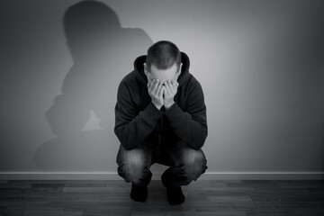 Young caucasian man crouched and holding his face. Concept photo of depression, anxienty, solitude and sorrow.