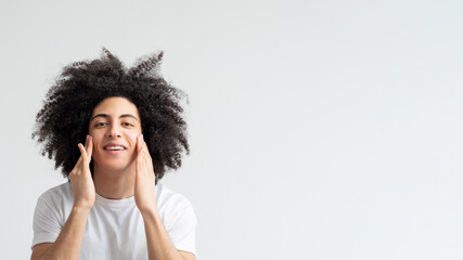 Man facial care. Skin treatment. Male grooming. Happy satisfied handsome guy with long brown curly hair in white t-shirt applying cream smiling isolated on neutral empty space advertising banner.