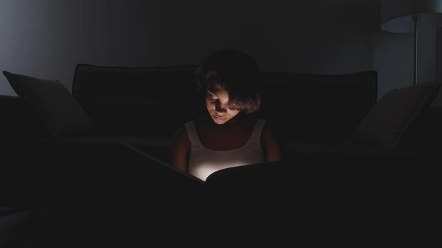 Portrait of young female sitting at home on the floor and reading a book, warm light, self-isolation during the pandemic, mystical lighting effect.