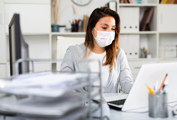 Woman in protective mask sitting at workplace with computer in her office