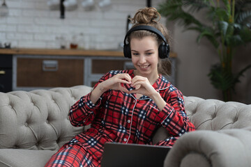 Girl teenager communicates on social networks or having video chat making a heart sign with his hands sitting on the couch at home