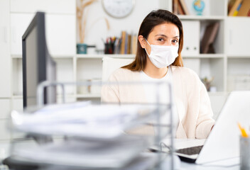 Fototapeta na wymiar Secretary girl in a protective mask during a pandemic sits at her workplace in the office in front of a laptop
