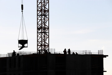 Silhouettes of workers and tower crane with cargo on construction site against the sky. Housing...
