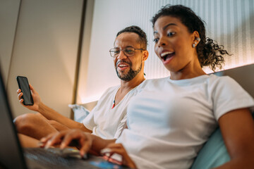 Happy Latinx couple sitting on bed, woman smiling using laptop