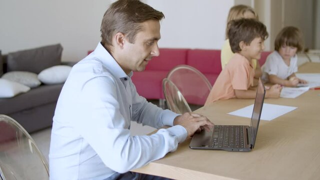 Caucasian dad working on laptop and cute kids painting doodles at table. Concentrated blonde girl drawing with marker and brother looking at her. Static camera. Childhood and weekend concept