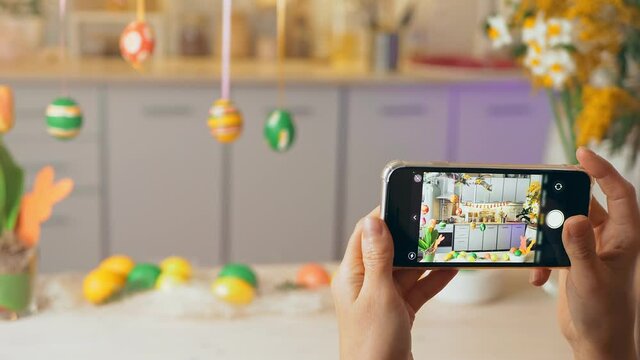 Hands hold smartphone. On display photo composition for holiday of spring Happy Easter. Girl takes pictures on phone painted eggs on Easter decorated on table of the house. 