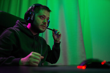 Gamer or streamer in headphones with microphone sits at home in neon light and plays at pc