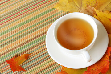 White cup with hot tea among the autumn leaves on a straw mat. Space for text on the left.