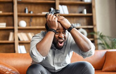 Front view portrait of young African American guy sitting on the couch in the living room, holding...