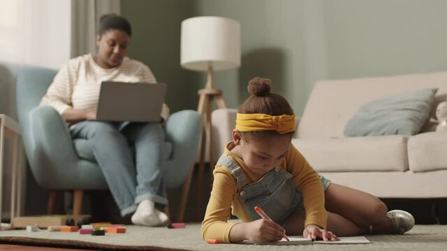 Wide shot of little African-American girl lying on carpet in living room and drawing in notebook while her mom sitting on armchair and using laptop on background