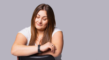 Banner- long format, gray background. Indoor image of charming positive overweight young Caucasian female relaxing, sitting on chair, having joyful facial expression. looking down