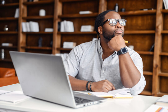 Serious thoughtful young African American man in glasses and hand on chin sitting at desk in office, looking away, taking break from working on laptop, pondering project plan, thinking of new ideas - Powered by Adobe