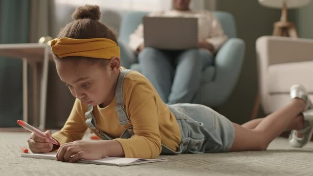 Lockdown of little African-American girl lying on carpet in living room and drawing in notebook using felt pen while her unrecognizable mom sitting on armchair with laptop on background