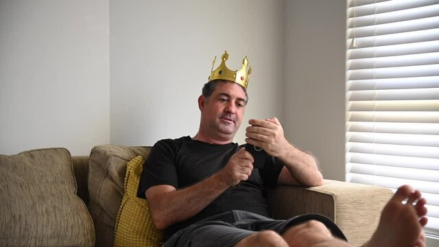 Lazy mature adult man (age 40-50) behaving like a king, lying on a couch looking at videos on mobile phone while his wife working and vacuuming living room. Laziness concept. Real people. Copy space
