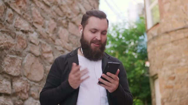 Low angle tracking shot of happy bearded man standing outside and talking on video call on mobile phone