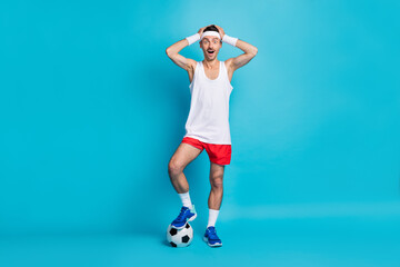 Full body portrait of impressed young man arms on head open mouth leg on ball isolated on blue color background