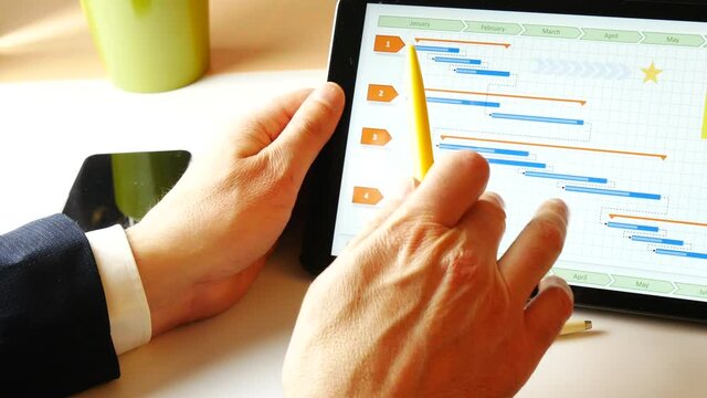 Business man scrolling tablet with a yellow pen, showing a gantt for project management purposes. Smartphone and pen on the desk