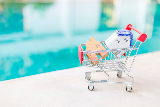 Miniature ceramic and wooden house model in shopping cart over blurred swimming pool background, summer sale, property business, outdoor day light