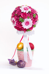 Easter composition with bright flower bouquet in pink pot, decorated by bands and bows, golden and red painted eggs. White background, copy space.