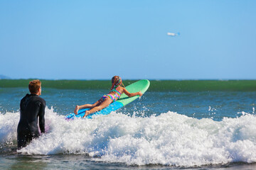 Little surf girl - young surfer learn to ride on surfboard with instructor at surfing school. Active family lifestyle, kids water sport lessons, swimming activity in summer camp. Vacation with child.