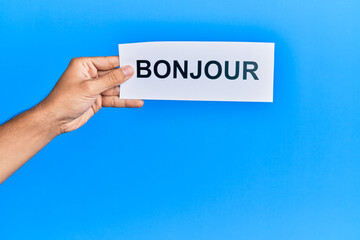 Hand of hispanic man holding french greeting bonjour word paper over isolated blue background. © Krakenimages.com