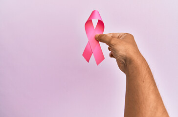 Hand of young hispanic man holding pink breast cancer ribbon over isolated background.