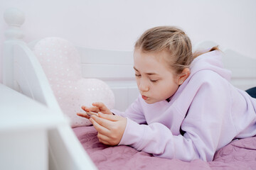 A Caucasian teenage girl uses a smartphone while resting on a bed in her room. The girl is video chatting with friends from school. Isolation. Remote learning. Coronavirus