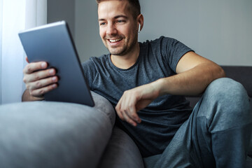 Fototapeta premium A middle-aged handsome man in the comfort and warmth of home in relaxed gray sportswear sits on a sofa in the living room of the apartment and looks at something on the tablet that makes him smile